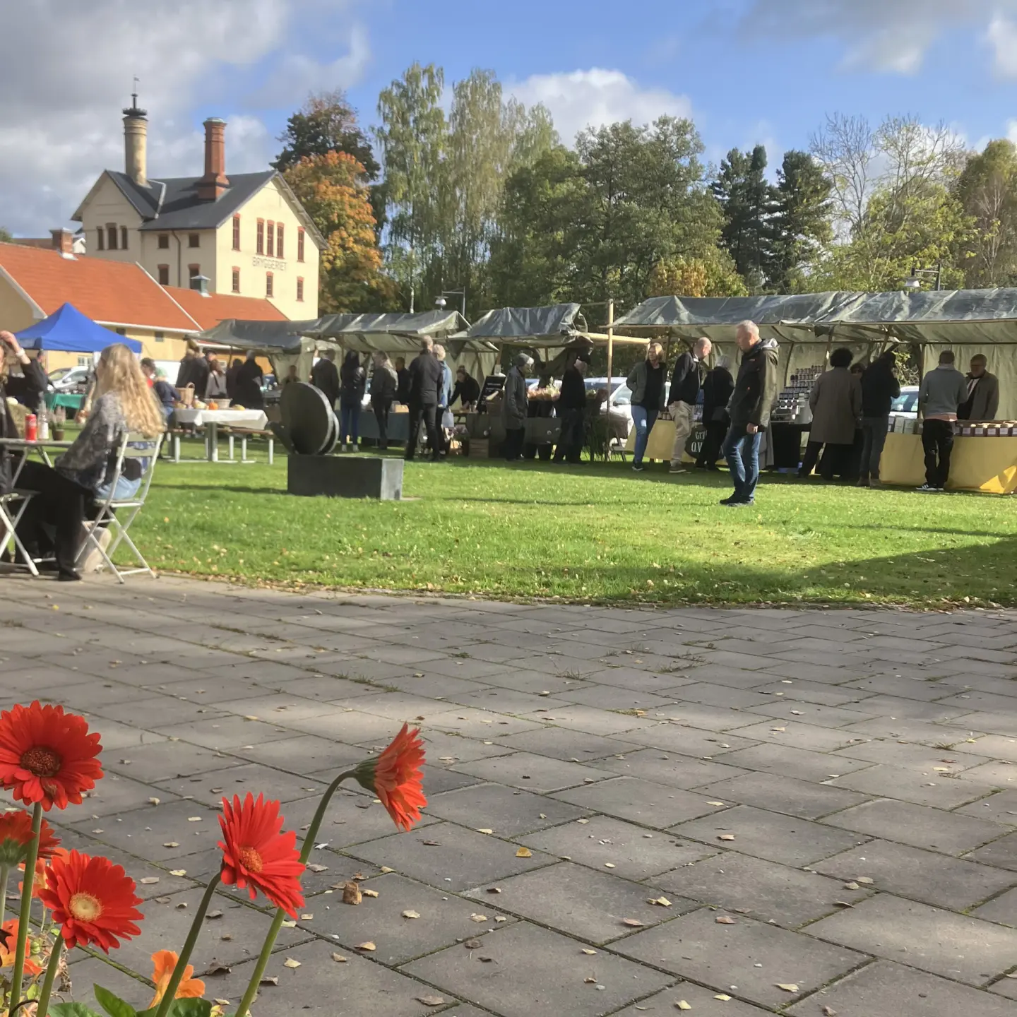 Höstmarknad i Kvarteret Bryggeriet med marknadsstånd och besökare på gräsmattan. I förgrunden syns röda blommor och i bakgrunden Bryggeriets byggnader och höstfärgade träd.