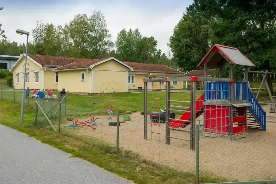 Bild på förskolan utifrån med utegård. Byggnaden är gul med rött tak. På bilden av gården finns det en sandlåda, klätterställning med rutschkana, gungställning och gräsmatta med staket runtomkring