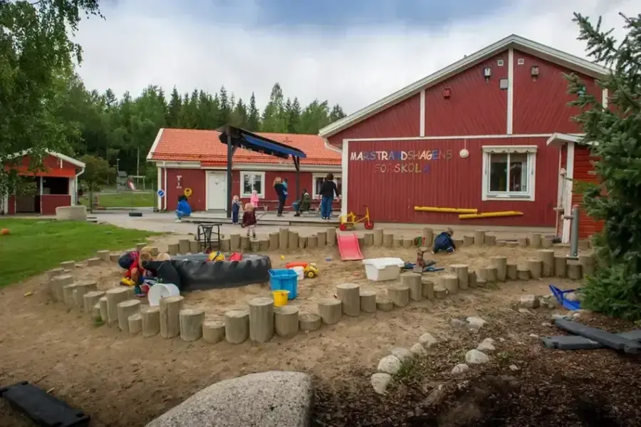 Bild utifrån på förskolan med utegård. Byggnaden är röd och har texten Marstrandshagens förskola på väggen. På gården finns en stor sandlåda med leksaker