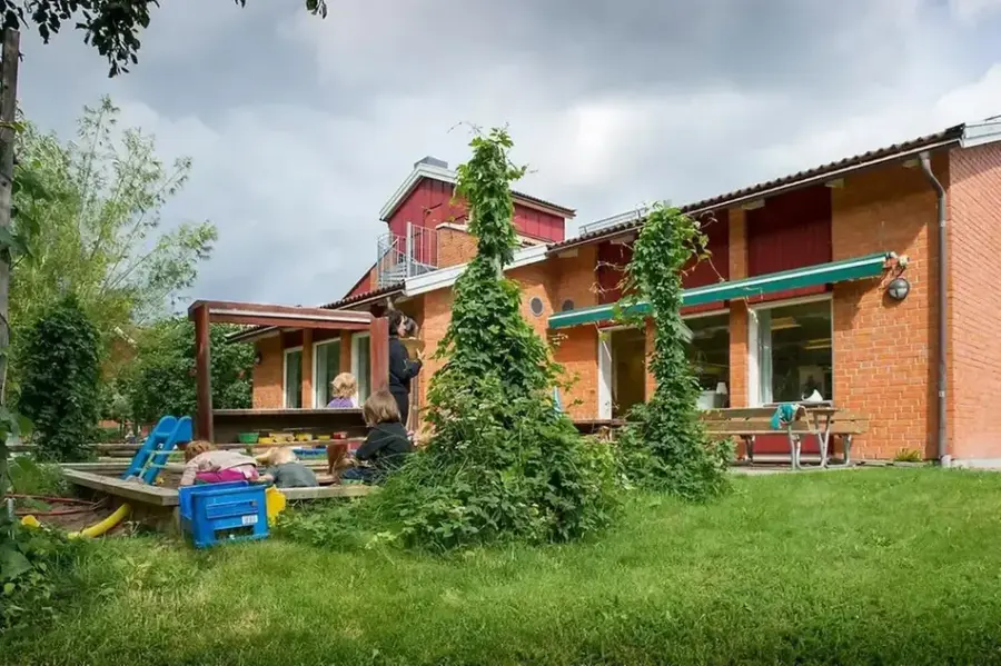 Bild på förskolan utifrån med utegård. Förskolan är en röd tegelbyggnad. Utegården har humleväxter och en sandlåda.