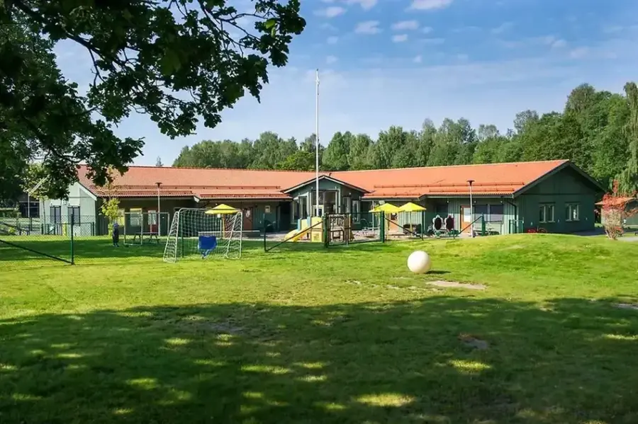 Bild utifrån på förskolan och utegård. Byggnaden är grön med orange tak. På gården finns det lekställningar, sandlåda och en gräsyta med fotbollsplan