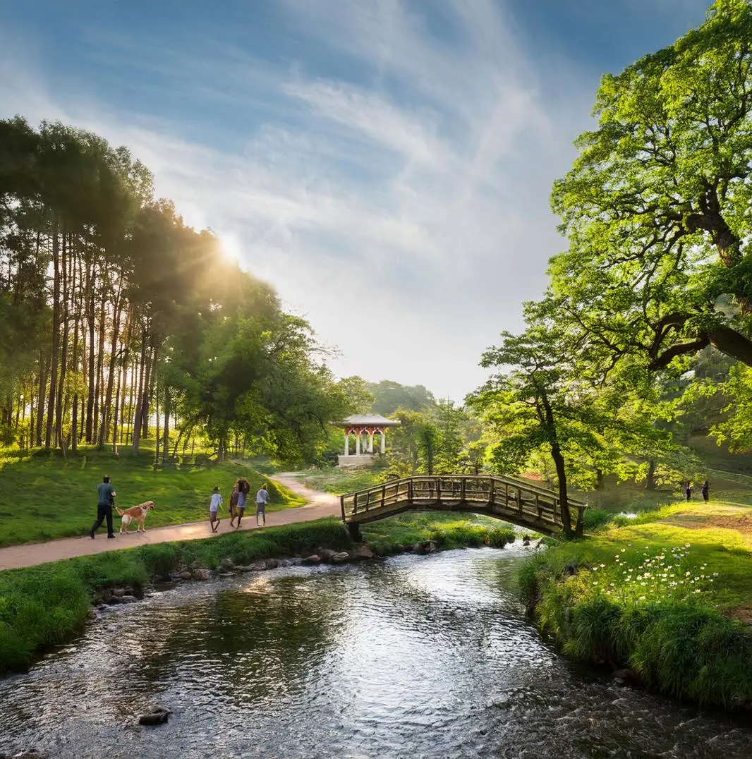 En AI-genererad bild av en vision hur det skulle kunna se ut vid hagbyparken. På bilden syns familjer och personer gå genom en grönskande park med vattendrag, paviljong och broar
