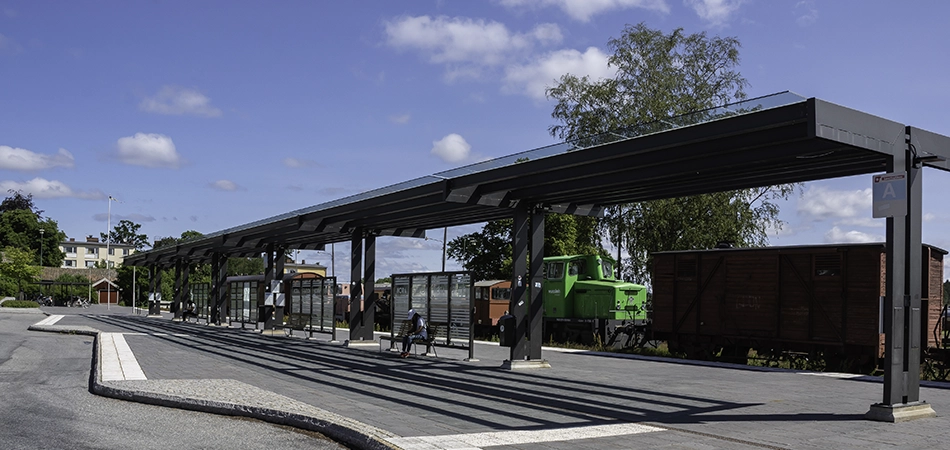 Stationsområdet, bilden visar busskurer med glastak och bänkar att sitta på, bakom syns järnvägen med flera vagnar och ett grönt diesellok