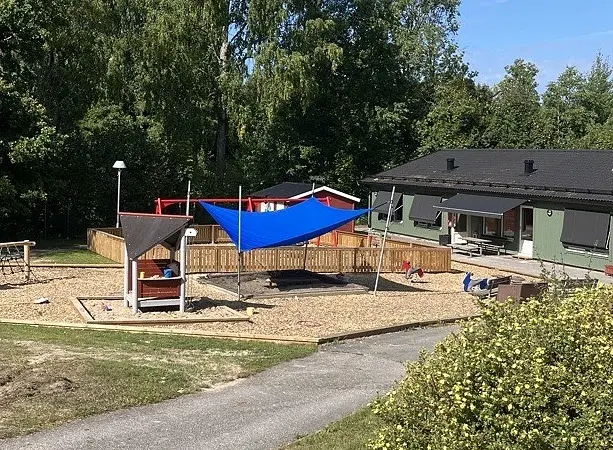Bild på förskolan utifrån med utegård. Byggnaden har grön fasad med svart tak. På utegården finns det lekställning, gungor och sandlåda med solskydd