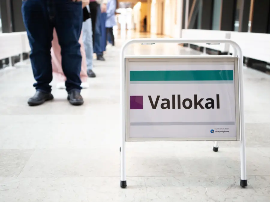 Fotografi av personer som står i kö, i förgrunden finns det en skylt där det står "vallokal"